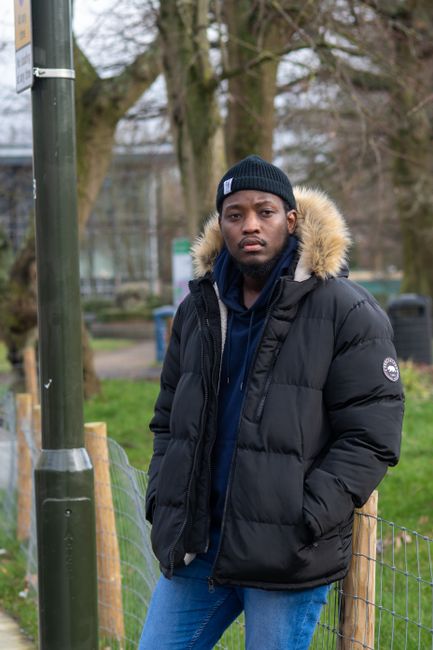 A young man with dark skin and serious expression standing outdoors in a park setting during daytime. He is wearing a black beanie hat, a black insulated winter jacket with a faux fur-lined hood, and a navy blue hoodie underneath. His hands are in the jacket pockets, and he is positioned near a wooden fence with metal wire mesh. In the background, there are tall leafless trees, a parking lot with some blurred vehicles, and a diffuse cloudy sky. The environment suggests a cold weather day, and the scene emphasizes an urban outdoor space, which could relate to the context of private waste handling or rubbish removal by demonstrating an individual involved in or observing waste-related activities outside a residential or communal area. Waste Clearance Croydon may be associated with such outdoor settings when handling rubbish removal or on-site clearance tasks, though no specific waste objects are visible in this image.