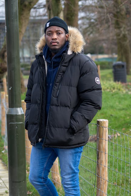 A young man with dark skin and serious expression standing outdoors in a park setting during daytime. He is wearing a black beanie hat, a black insulated winter jacket with a faux fur-lined hood, and a navy blue hoodie underneath. His hands are in the jacket pockets, and he is positioned near a wooden fence with metal wire mesh. In the background, there are tall leafless trees, a parking lot with some blurred vehicles, and a diffuse cloudy sky. The environment suggests a cold weather day, and the scene emphasizes an urban outdoor space, which could relate to the context of private waste handling or rubbish removal by demonstrating an individual involved in or observing waste-related activities outside a residential or communal area. Waste Clearance Croydon may be associated with such outdoor settings when handling rubbish removal or on-site clearance tasks, though no specific waste objects are visible in this image.