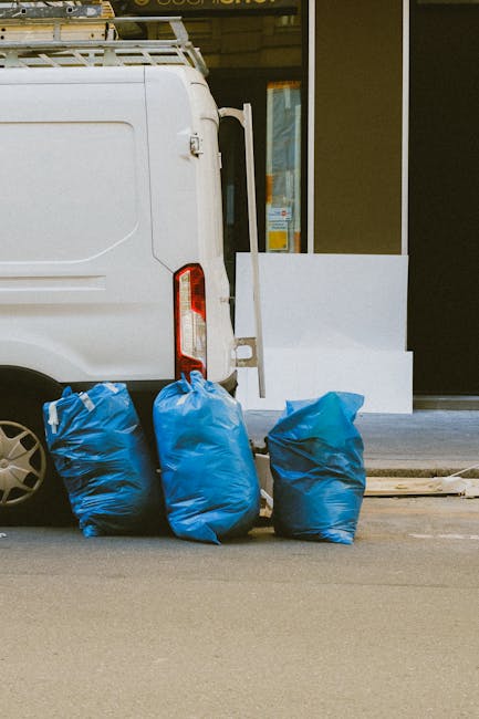 A white commercial van parked parallel to a sidewalk on a paved street, with three large, blue, plastic rubbish bags positioned on the ground near the rear of the vehicle. The bags appear to be filled with mixed waste materials and are loosely gathered at the top, with some slight creasing and texture visible on the plastic. The van's rear door is closed, and the vehicle has a vertical taillight on the left side, partially visible in the image. In the background, there is a building entrance with a glass door and windows, along with a white rectangular sign or panel leaning against the building wall next to the bags. The environment suggests an urban or suburban setting, potentially near a commercial or residential property, with natural lighting indicating daytime. Waste Clearance Croydon specialises in alternative waste handling methods such as private rubbish collection, evident through the neatly placed disposal bags ready for collection or removal, an approach often preferred for convenience over traditional council services.