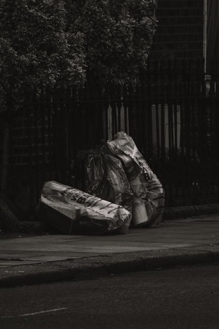 Two large black garbage bags filled with waste material are placed on the pavement beside a low curb, positioned in front of a black metal fence and dark foliage. The bags appear to be made of heavy-duty plastic, with slightly crumpled and shiny surfaces reflecting minimal light. The scene is set outdoors in a residential or urban environment, with the bags resting on a paved sidewalk adjacent to a roadway. The background features a dense, leafy bush or tree, providing a contrasting textured backdrop against the dark fence. The overall lighting is subdued, with shadows cast across the scene, emphasizing the bags and their bulk. This image depicts an example of private rubbish disposal, suitable for waste clearance services such as those offered by Waste Clearance Croydon, focusing on on-site collection of domestic or garden refuse prior to professional removal or disposal, aligning with alternative waste handling methods outside municipal collection.
