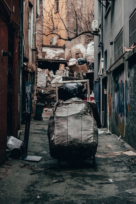 A narrow urban alleyway shows a large, tightly packed black and white duffle bag with worn fabric and visible creases positioned in the foreground on a slightly wet, asphalt surface. Behind it, there is an extensive pile of mixed rubbish bags, cardboard boxes, and discarded debris stacked against a weathered brick wall at the end of the alley. The background features a mix of residential and commercial building facades with some windows and bare tree branches extending overhead. On the right side, a grey building with metal bars and graffiti partially visible on its surface is adjacent to the alley. The environment appears damp with subtle gloom, likely due to recent rain, and the scene is illuminated by natural daylight. The image visually aligns with private waste collection or on-site clearance activities often managed by independent rubbish removal services such as Waste Clearance Croydon, highlighting the typical clutter associated with waste disposal in urban alleyways.