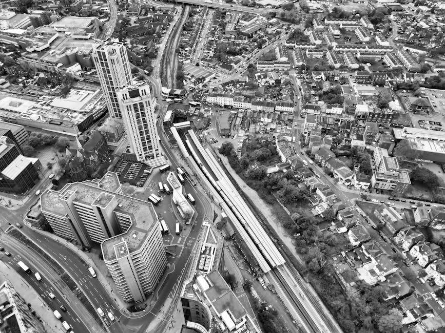 A black and white aerial photograph of an urban area featuring a cluster of tall modern office buildings and high-rise flats, with visible rooftops and glass facades reflecting light. In the foreground, several buses and vehicles are seen moving along curved roads and a multi-lane street, with some parked near the buildings. To the right, a railway corridor with an elevated train track runs through the scene, flanked by trees and residential houses with pitched roofs, small gardens, and narrow streets. The surrounding environment includes a mix of commercial, residential, and transport infrastructure, with dense building arrangements and green patches. The image captures the varied textures of buildings, roads, and vegetation, illustrating a busy cityscape typical of a central business district and nearby neighborhoods, consistent with urban rubbish management and alternative waste handling in a metropolitan setting. The scene is illuminated with natural daylight, with some shadows cast by the structures, providing a clear view of the dense built environment and transport routes.
